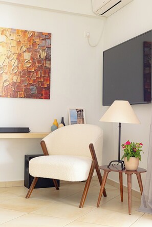 Ein modernes Zimmer mit einem weißen Sessel, einem kleinen Beistelltisch mit Lampe und Topfpflanze, einem großen abstrakten Gemälde und einem flachbildschirmfernseher an der Wand.