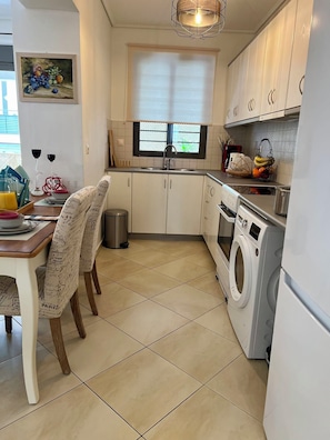 A compact kitchen with a washing machine, a dining table with chairs, and a window with blinds. - Elegant Apt 50meters Close Acropolis (Athina)