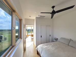 Quarto com cama, ventilador de teto, vista pela janela grande e porta que dá para uma varanda.