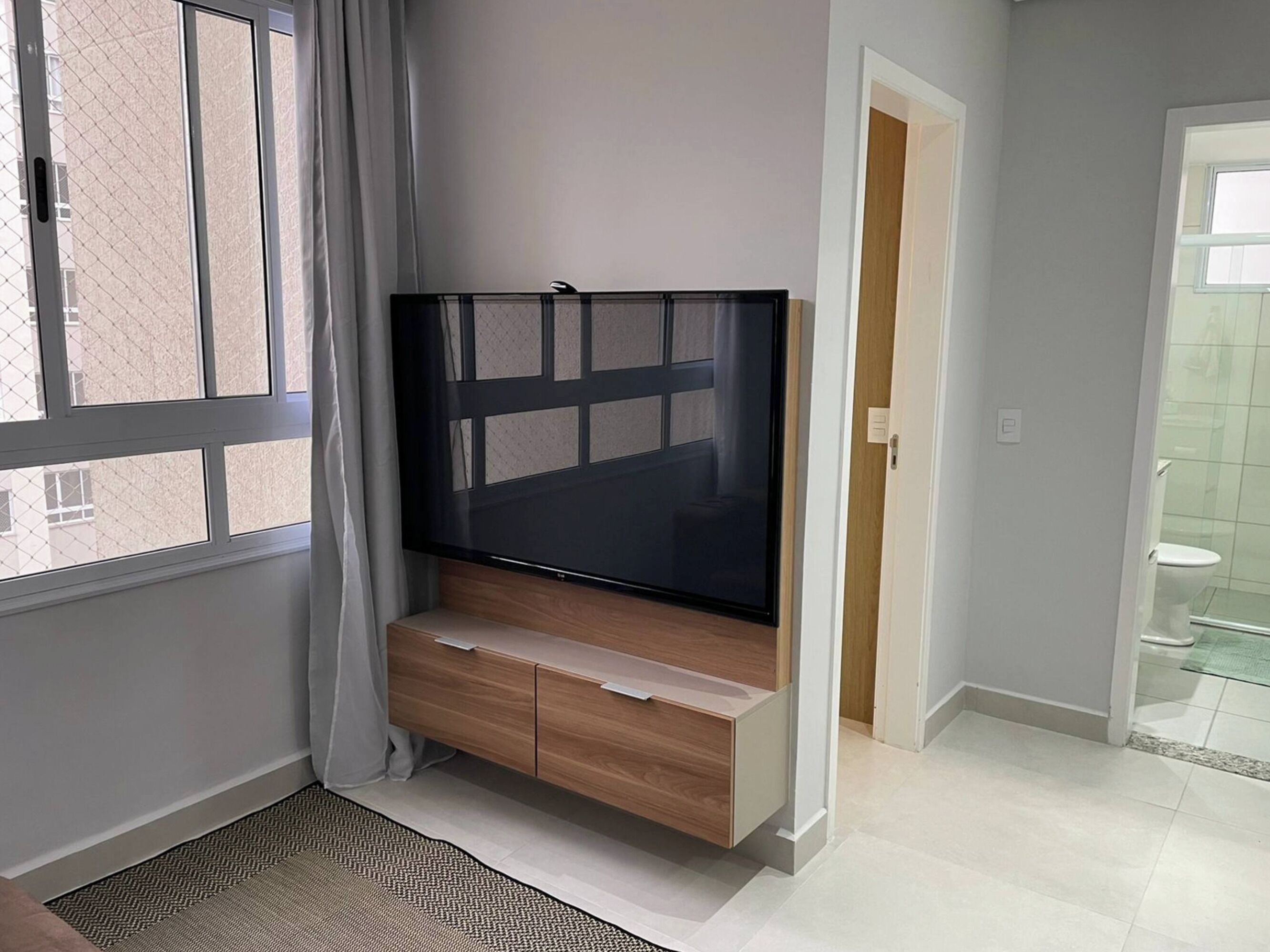 Sala de estar moderna com TV de tela plana em suporte de madeira, uma janela grande com cortinas e um banheiro visível através de uma porta aberta.