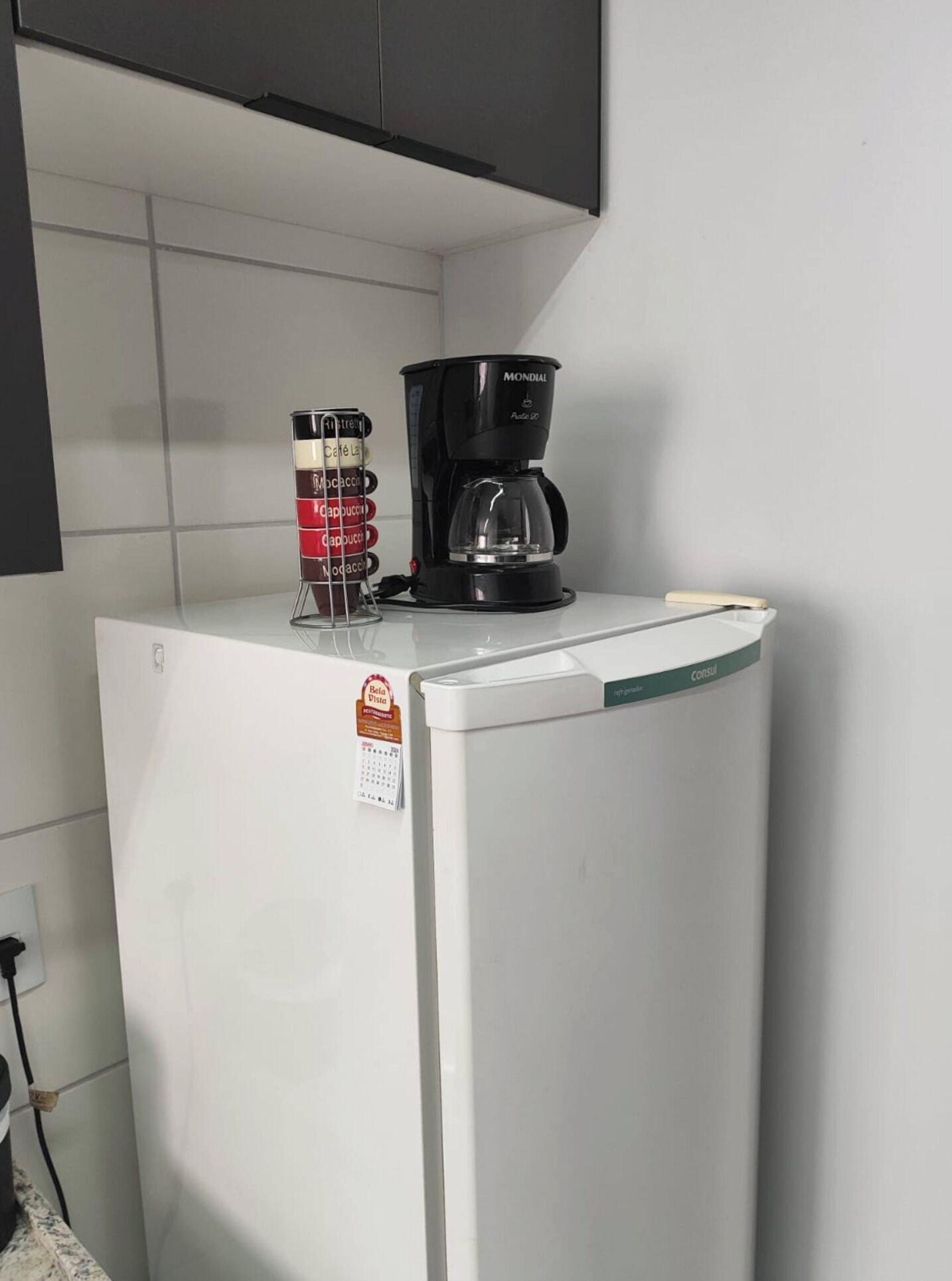 Un réfrigérateur blanc, avec une machine à café posée sur le dessus, un calendrier et un récipient doté d’étiquettes.