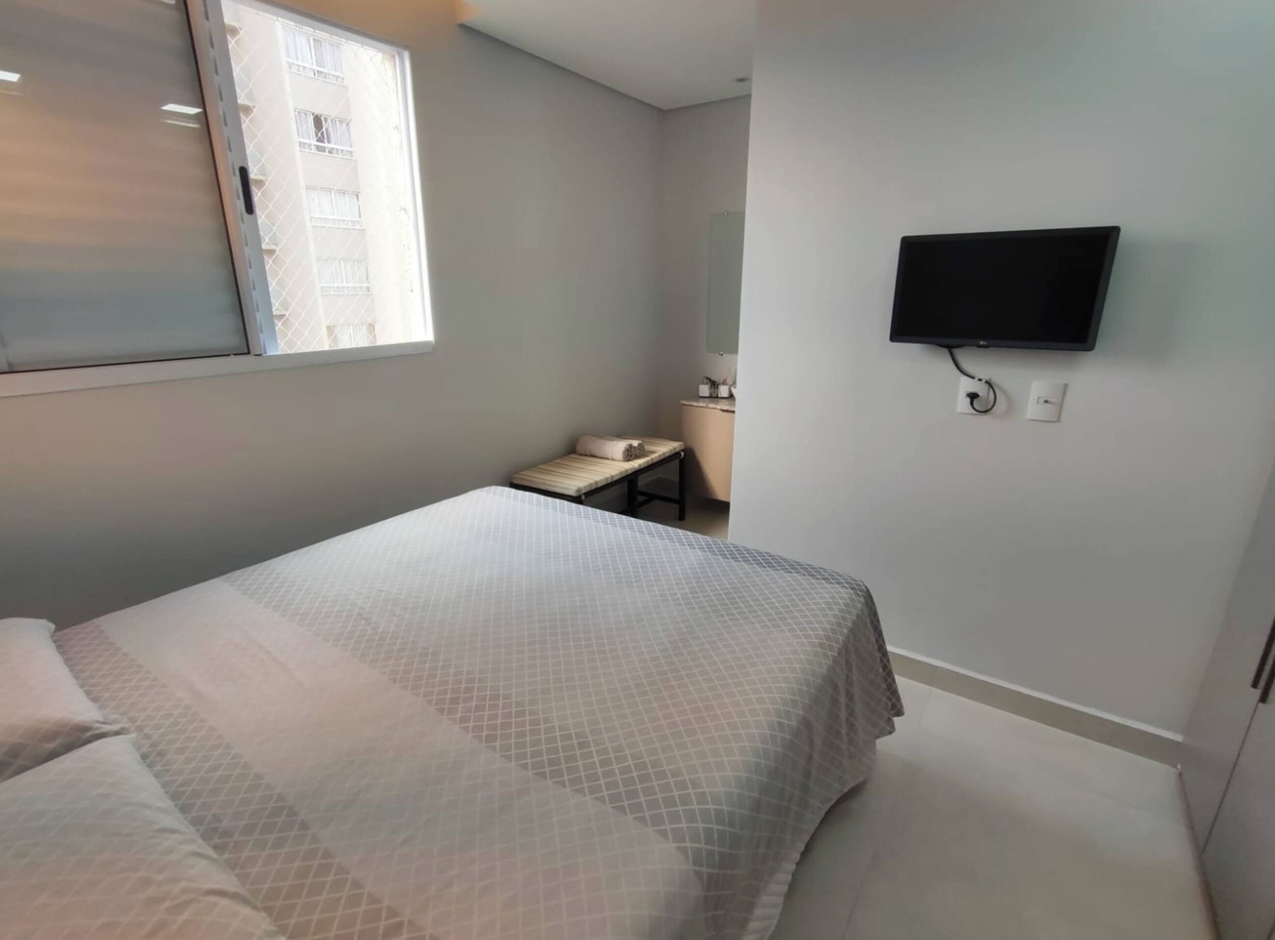 Una habitación de hotel con cama, televisor montado en la pared, ventana con persianas y una mesita de noche con lámpara.