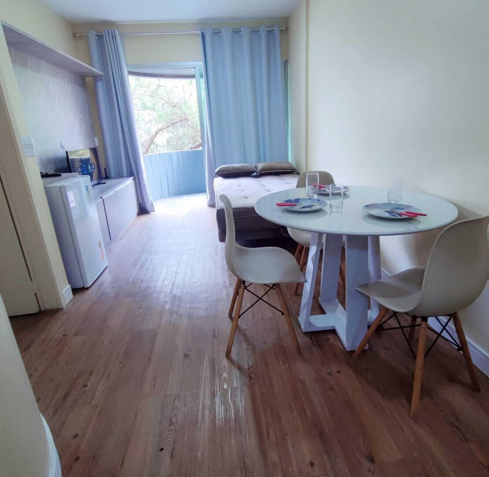 A room with a dining table, chairs, a kitchenette, a bed, and a window with curtains.