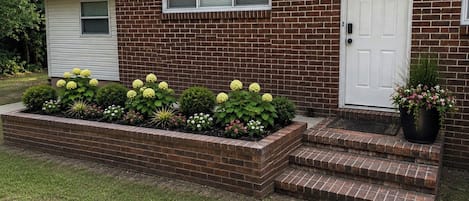 Una casa de ladrillo de dos pisos con una puerta frontal blanca, un ventanal pequeño y un parterre con diversas plantas.