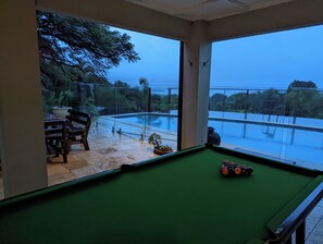 Ein Billardtisch mit Kugeln, ein gläserner Bereich mit Blick auf eine Brücke und Grünflächen sowie eine Sitzecke mit Tisch und Stühlen.