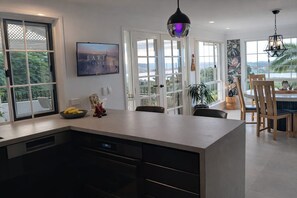 A modern kitchen with a large island, a dining area with wooden chairs, and a view of the outdoors. - Large, modern, resort style property with tropical gardens, pool, volleyball. (Terranora)