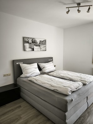 A modern bedroom with a grey bed, two pillows, a headboard, a bedside table, a nightstand, a wall-mounted light fixture, and a small picture frame.