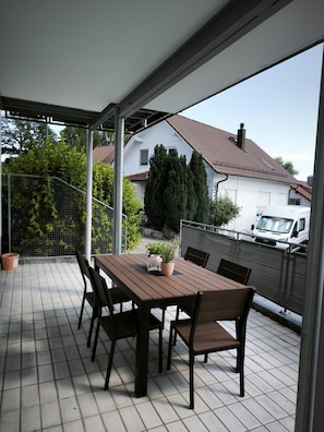 A covered outdoor patio with a wooden table and chairs, a potted plant, and a view of a white house and parked vehicle.