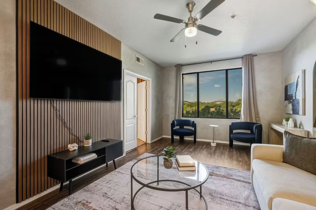 A modern living room with a flat-screen TV, a ceiling fan, a large window with a view, and a sofa.