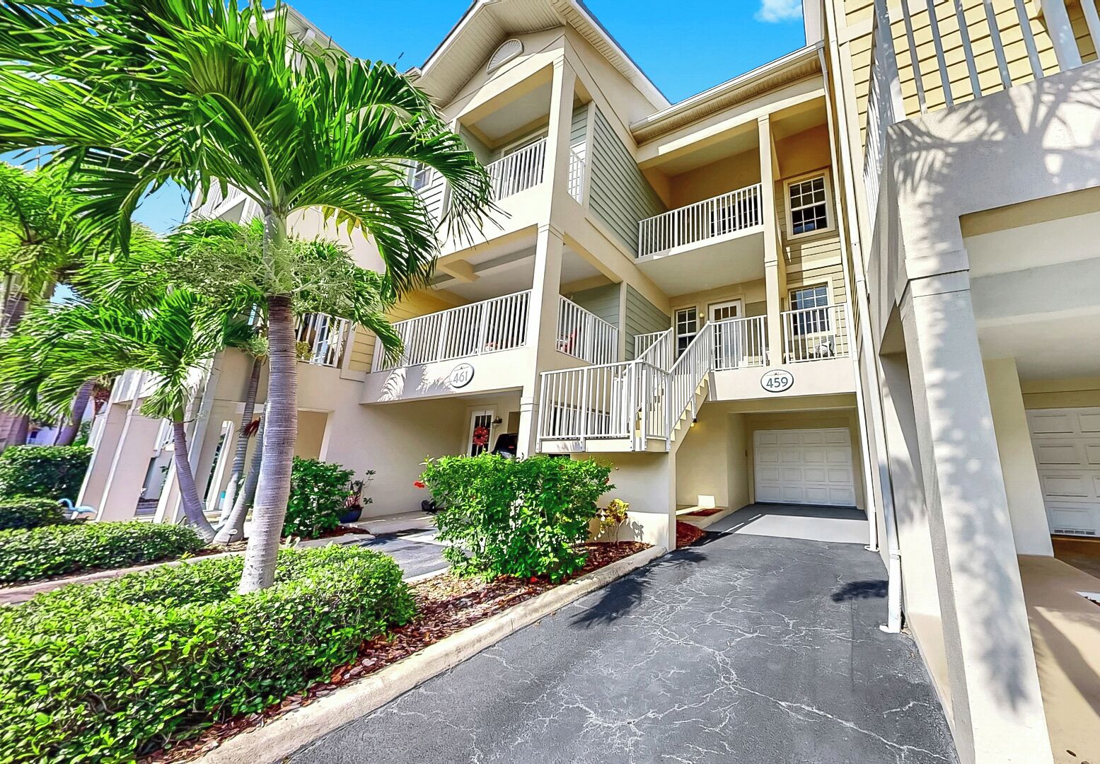 A multi-story residential building with balconies, a garage, and a well-maintained garden with palm trees and shrubs.