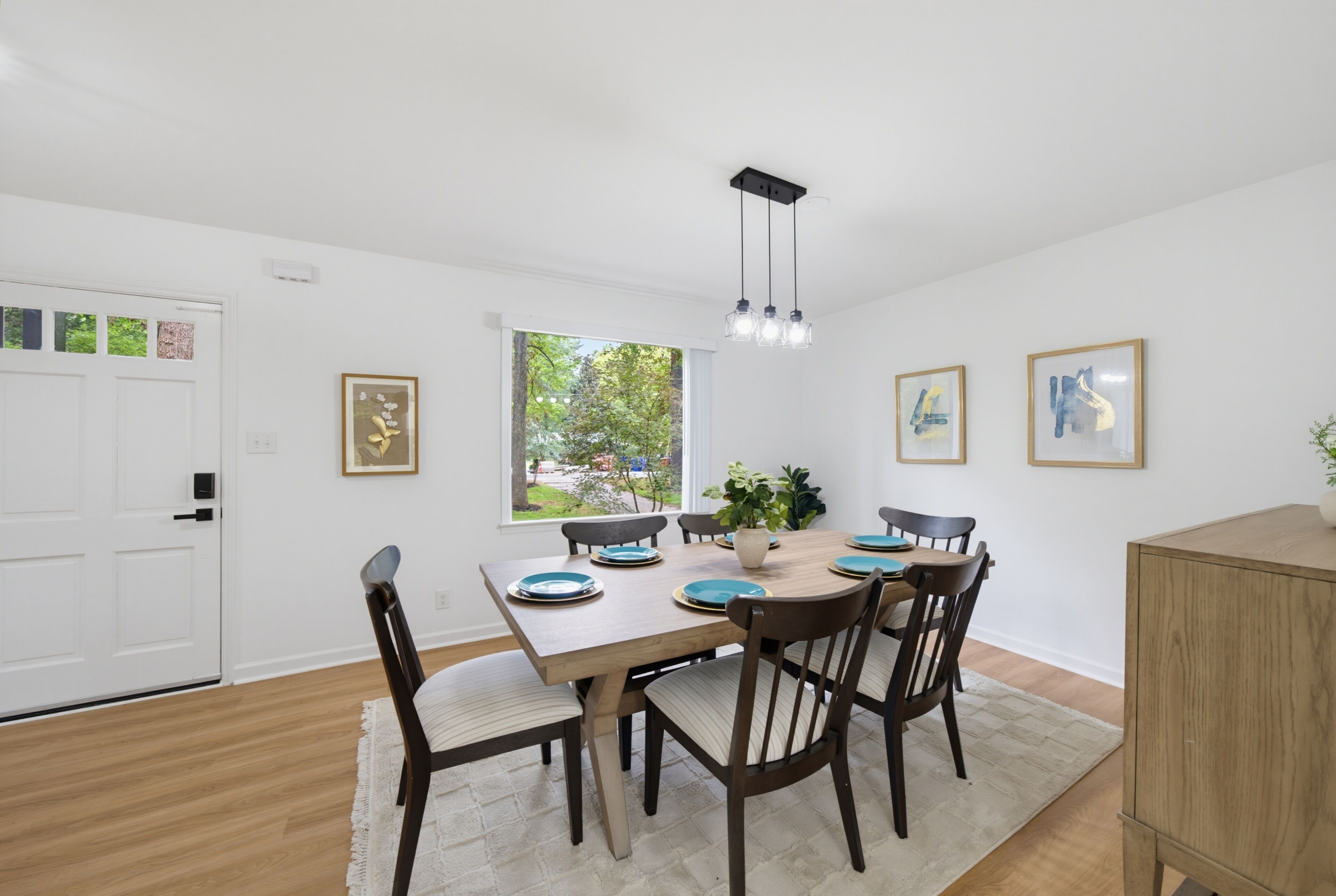 A dining area with a wooden table, chairs, and a chandelier. There are framed pictures on the walls and a window with a view of greenery.