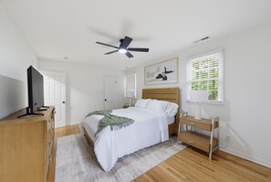 Une chambre à coucher comprenant un lit, un bureau, une télévision, un ventilateur de plafond, une fenêtre avec des stores et un tableau encadré accroché au mur.