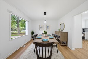 A dining room with a wooden table set for four, flanked by chairs, a rug, and a view of trees through a large window.