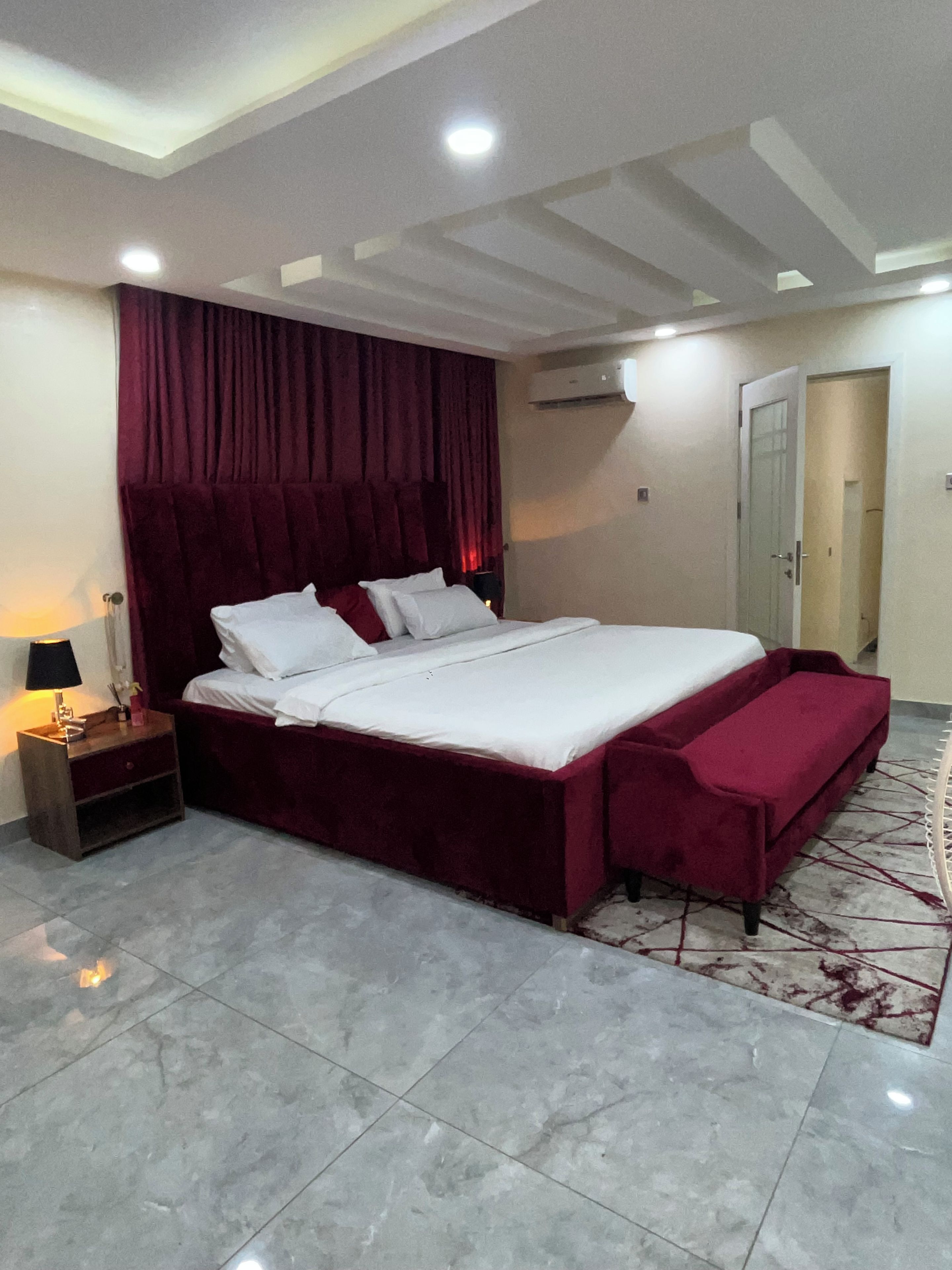 Une chambre à coucher avec un grand lit, une table de chevet avec une lampe, un rideau rouge, un appareil de climatisation et une porte.