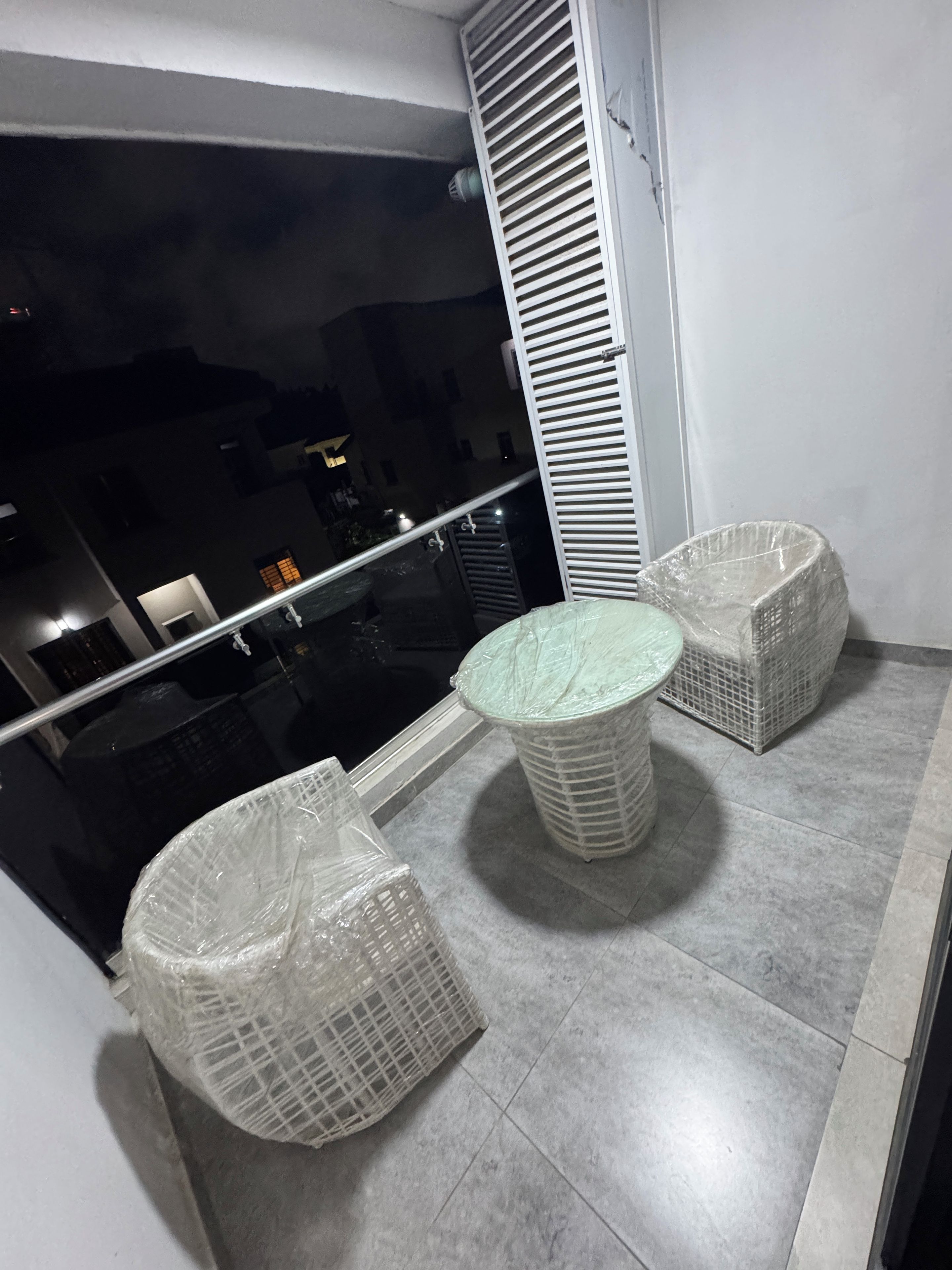 Un balcón con barandilla de vidrio, una mesa redonda y dos sillas de mimbre.