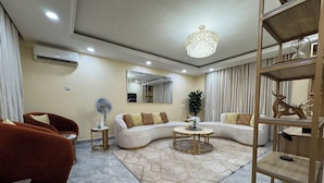 A modern living room with a sectional sofa, a round coffee table, a floor lamp, and a decorative shelf. - 4 Bed Luxury Town House (Lagos)