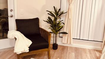 A living room with a dark-colored sofa, a potted plant, a round wooden coffee table with a remote control and decorative items, and a door with a towel on the armrest.