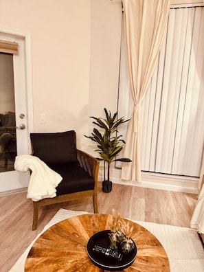Een woonkamer met een donkergrijze bank, een potplant, een ronde houten salontafel met een afstandsbediening en decoratieve objecten, en een deur met een handdoek op de armleuning.