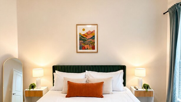 Una camera d'albergo con un letto grande, due comodini con lampade, una ventola a soffitto, un quadro appeso al muro e una finestra con tende.