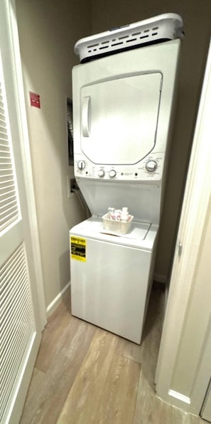 Een compacte, witte wasmachine met een mand vol wasgoed erop, geplaatst in een smalle gang met een houten vloer.