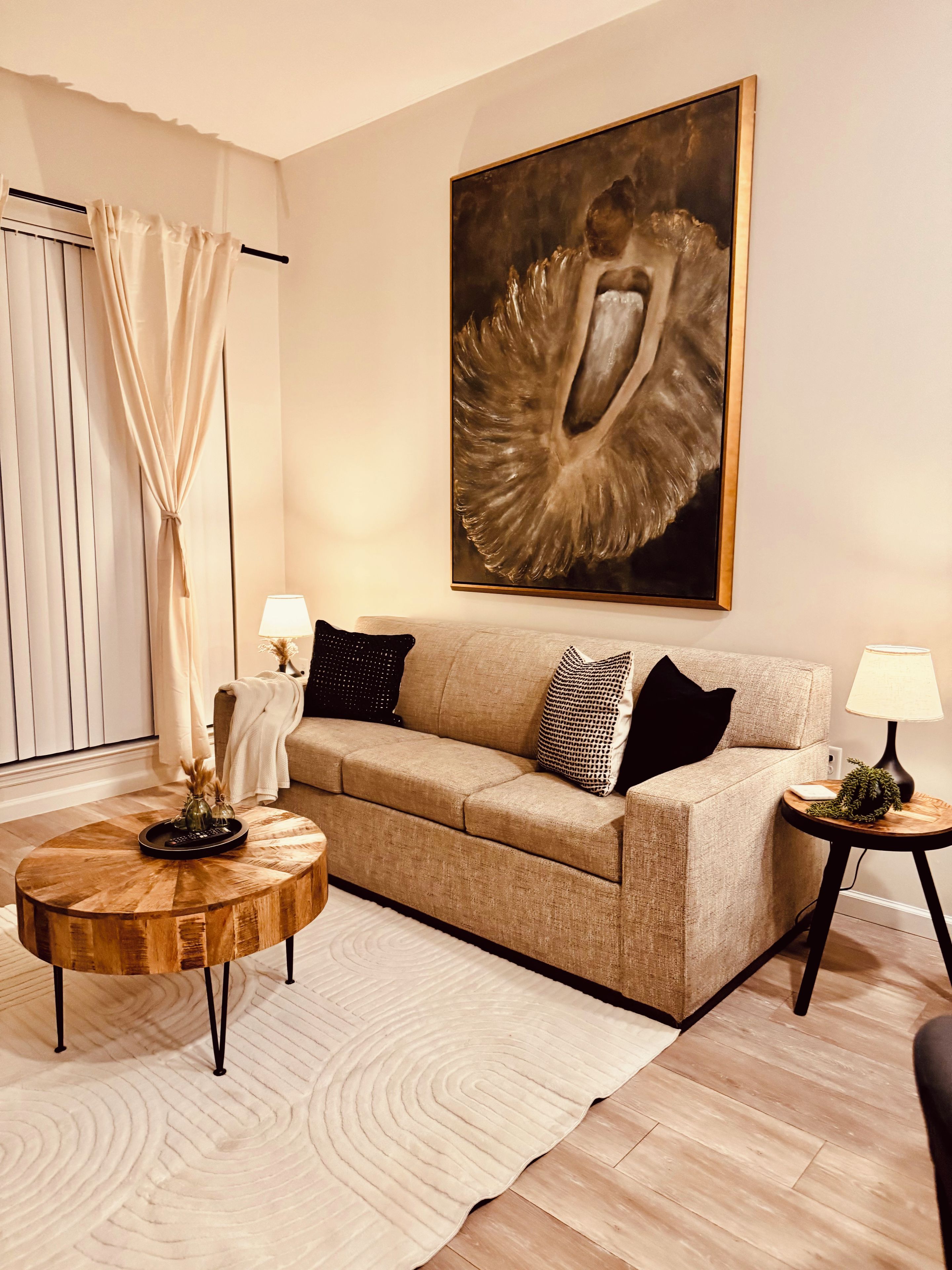 A living room with a beige sofa, a round coffee table, a painting on the wall, and a window with curtains.
