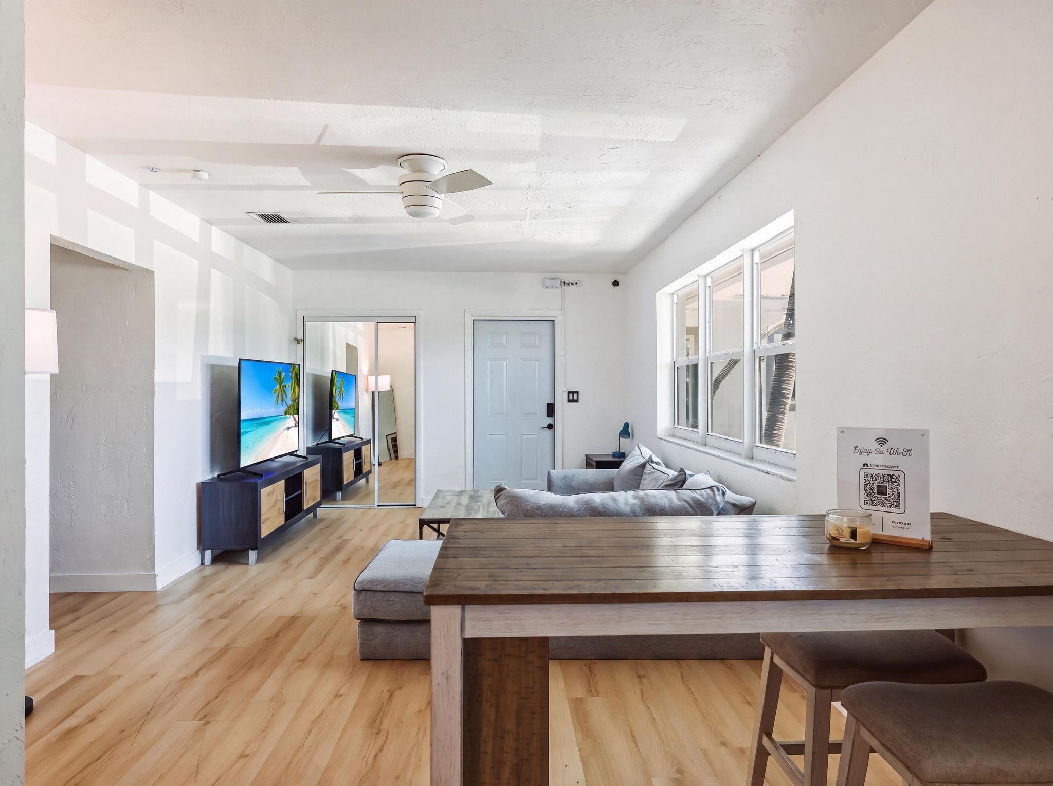 A modern living room with a wooden dining table, a sofa, and a TV displaying a beach scene.