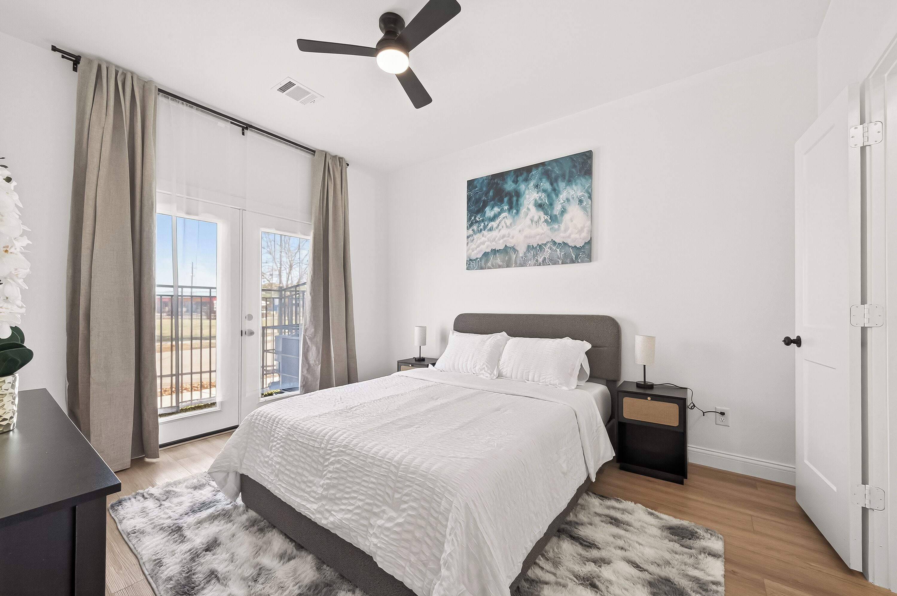 A bedroom with a bed, a ceiling fan, a painting, a nightstand, and a view through a window.