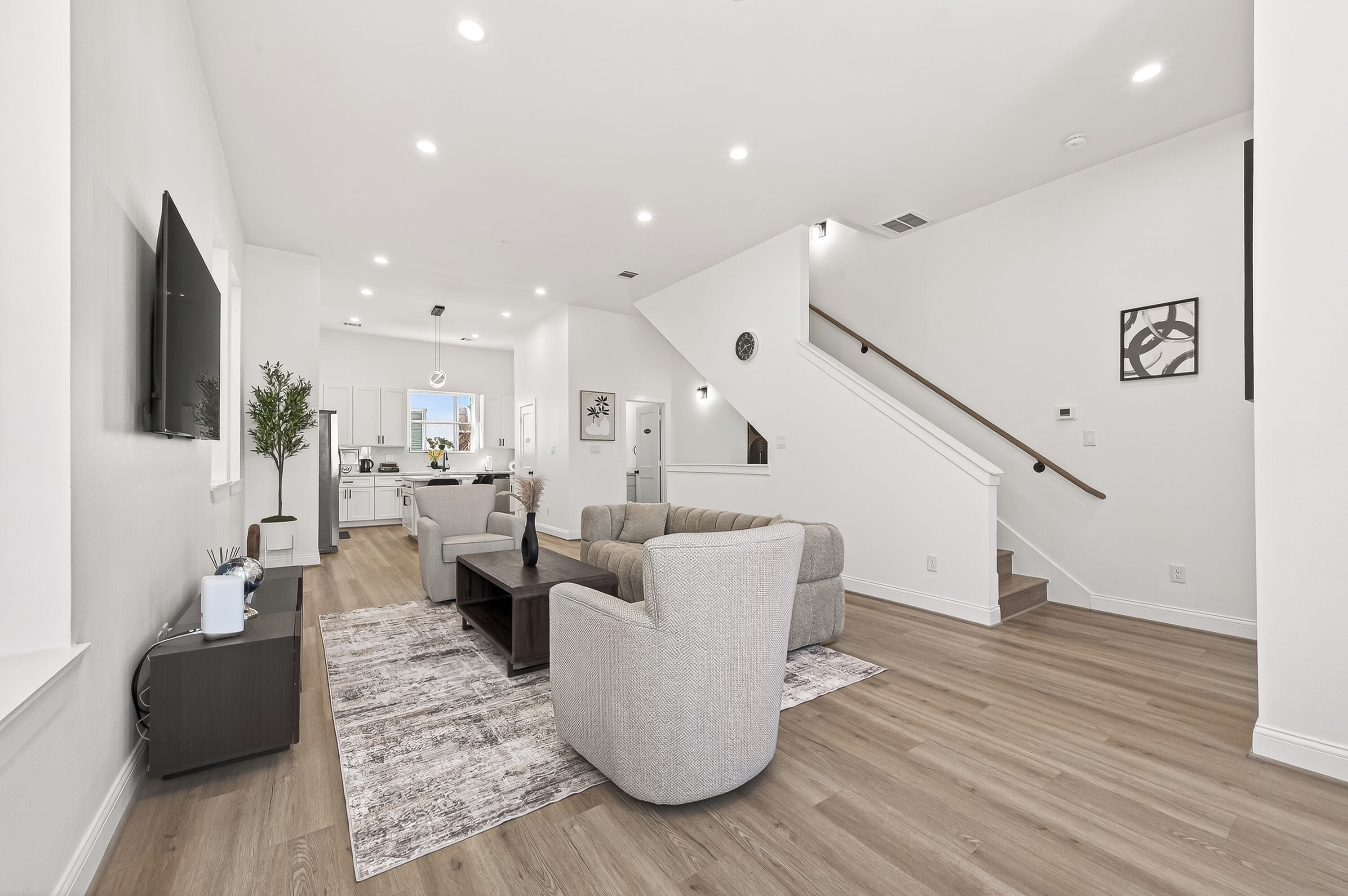 A spacious living area with a modern kitchen in the background, a staircase, and a flat-screen TV mounted on the wall.