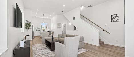 A spacious living area with a modern kitchen in the background, a staircase, and a flat-screen TV mounted on the wall.