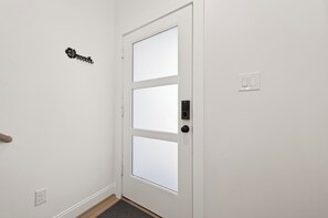 Eine weiße Tür mit einem mattierten Glasfeld, einem schwarzen Klingelknopf und einem Lichtschalter an der Wand.