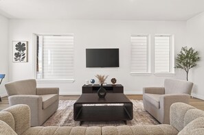 Ein Wohnzimmer mit Flachbildfernseher an der Wand, zwei Sesseln, einem Couchtisch und einer Topfpflanze.
