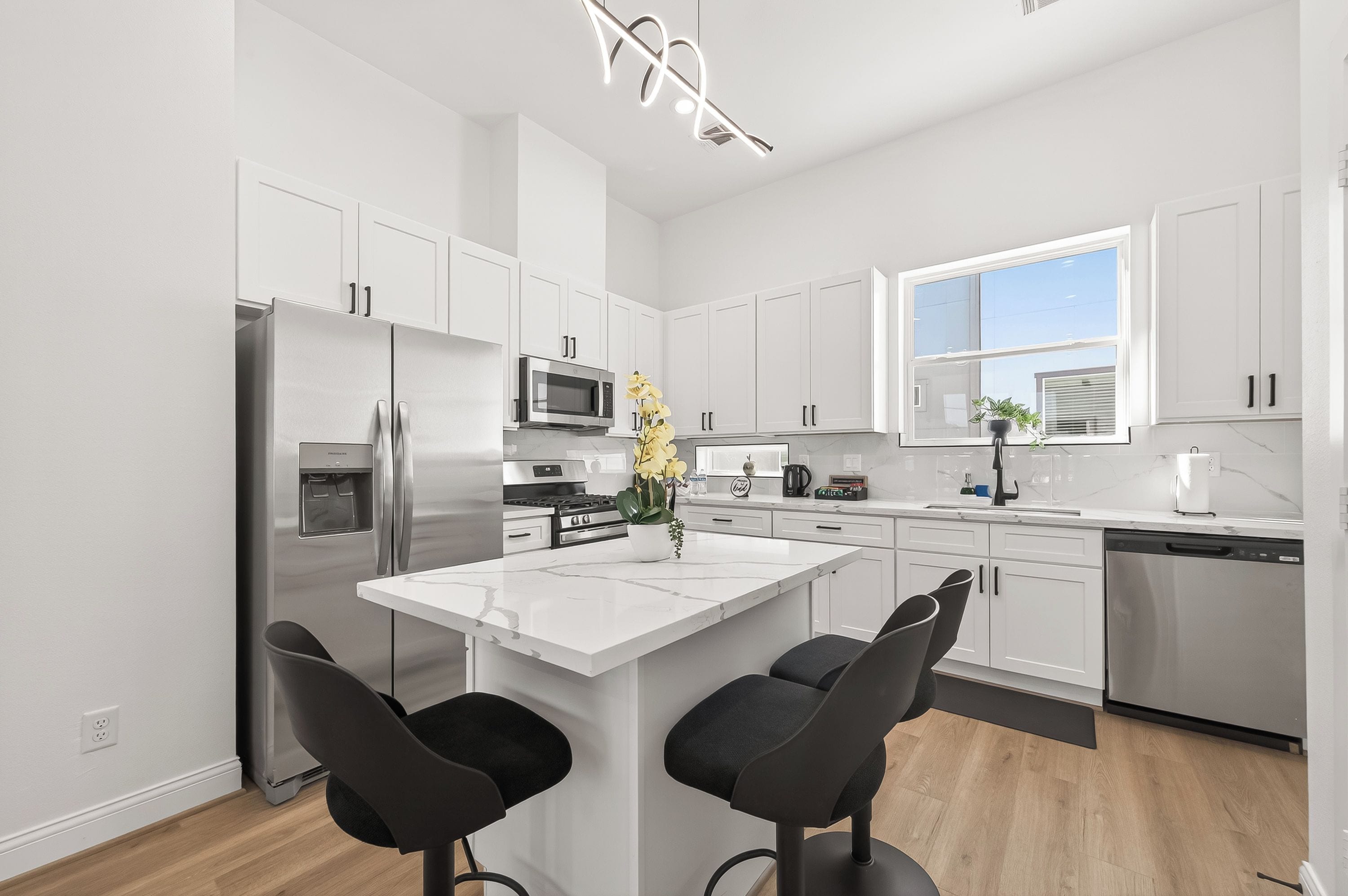 A modern kitchen with white cabinetry, stainless steel appliances, a marble countertop, and black bar stools.