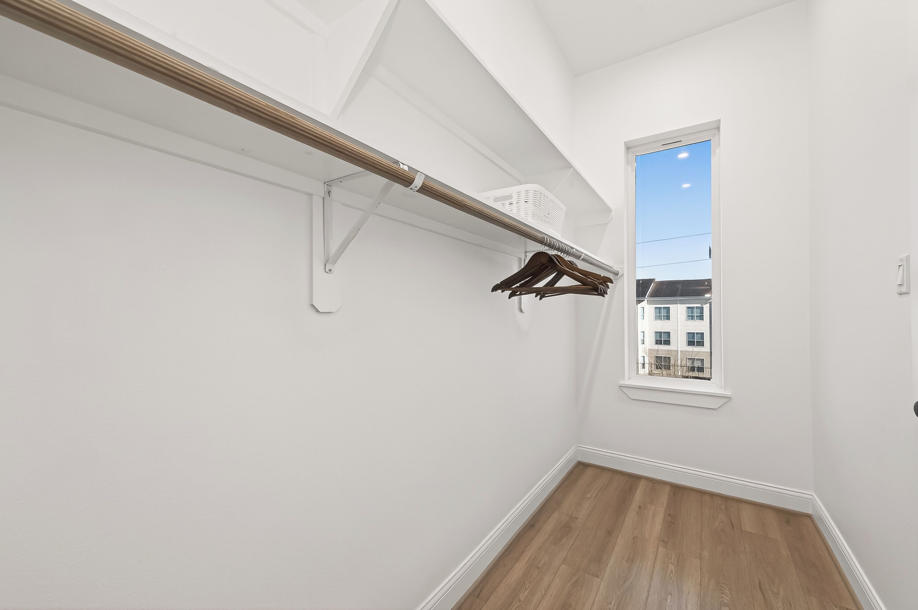 A hallway with a wooden floor, a white wall, a window with a view of buildings, and a clothes rack with hangers.