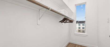 A hallway with a wooden floor, a white wall, a window with a view of buildings, and a clothes rack with hangers.