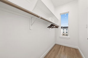 Ein Flur mit Holzboden, einer weißen Wand, einem Fenster mit Blick auf Gebäude und einem Kleiderständer mit Kleiderbügeln.