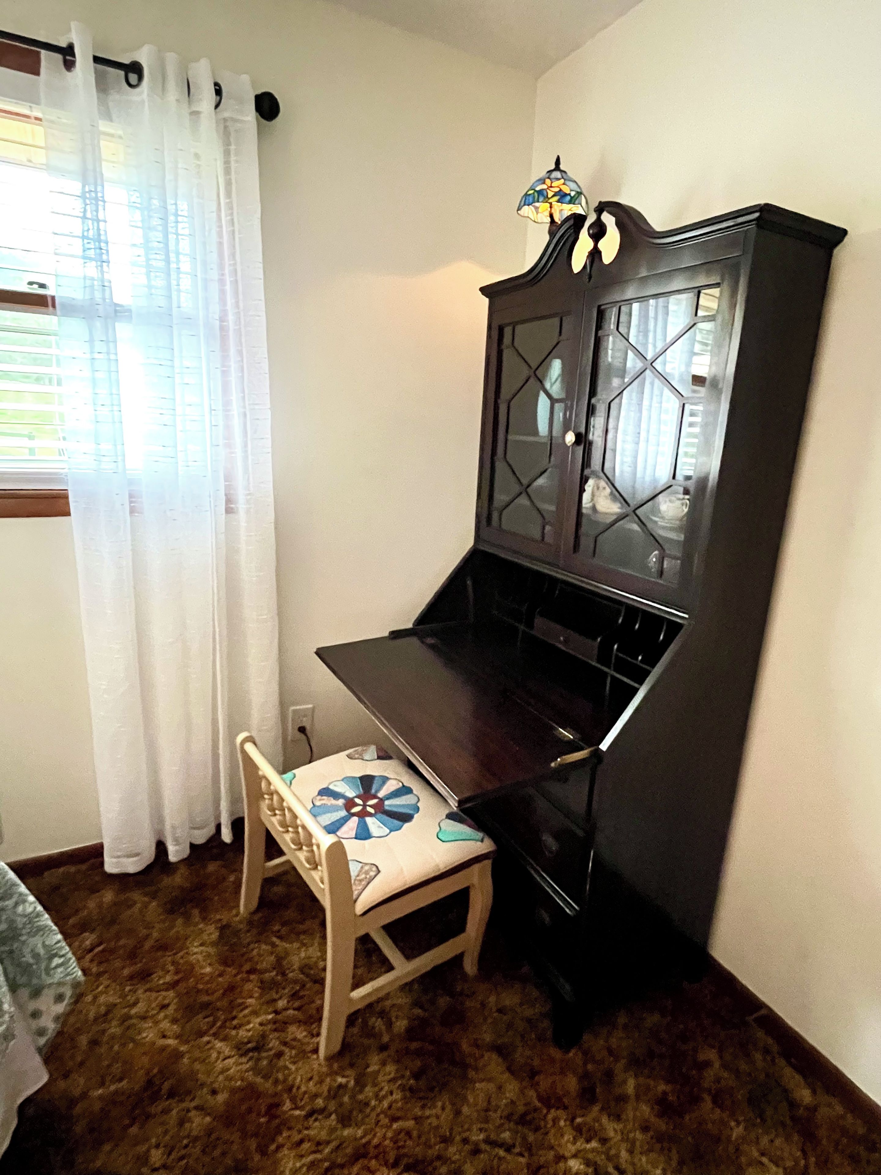 Una habitación de esquina con un piano, un aparador con puertas de cristal, una silla con cojín estampado y una ventana con cortinas translúcidas.
