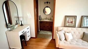 Une pièce avec une coiffeuse blanche, un miroir, un canapé blanc et une porte en bois donnant sur une salle de bain.