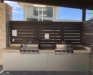 An outdoor grill station with two gas grills, a stone wall, and a wooden fence. - Accessible Queen Studio w/ Free Hot Breakfast (Saskatoon)