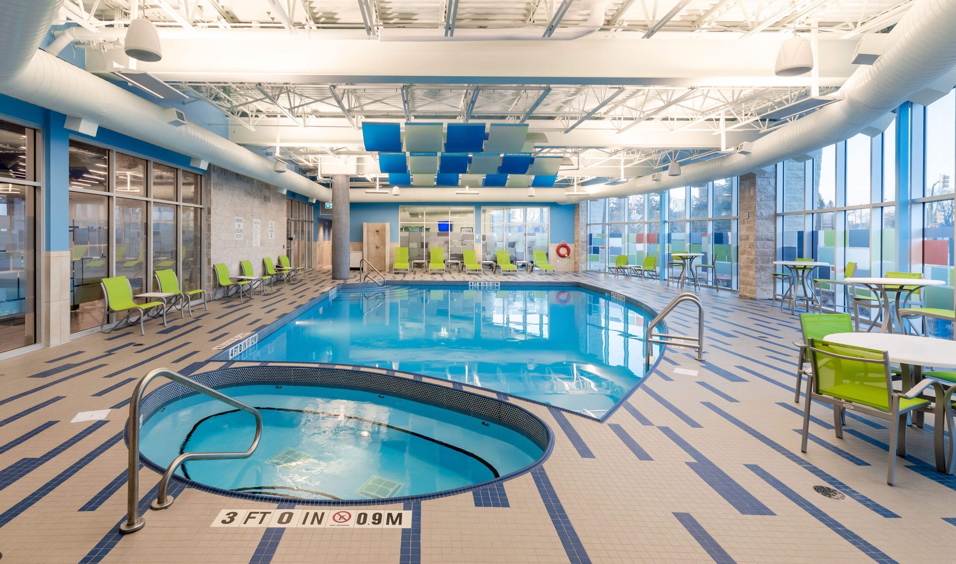 An indoor swimming pool with lounge chairs and tables, large windows, and a tiled floor.