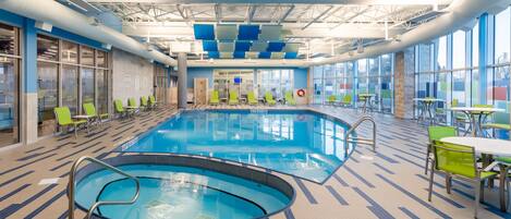 An indoor swimming pool with lounge chairs and tables, large windows, and a tiled floor.