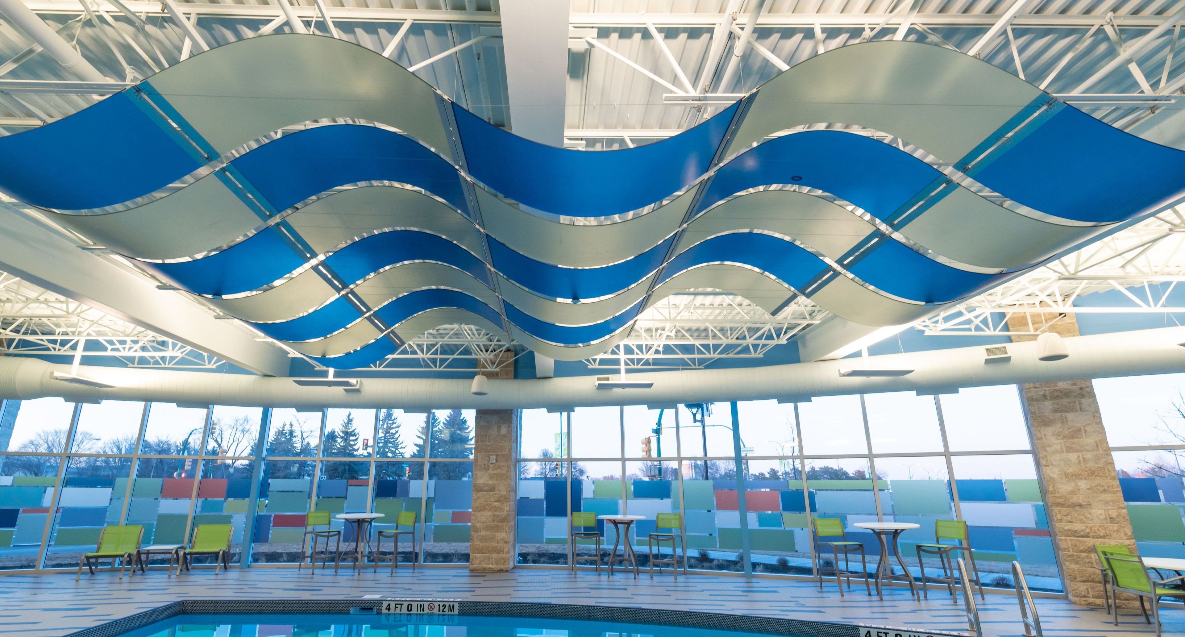 An indoor swimming pool with a wave-like ceiling design, clear glass walls, and multiple seating areas.