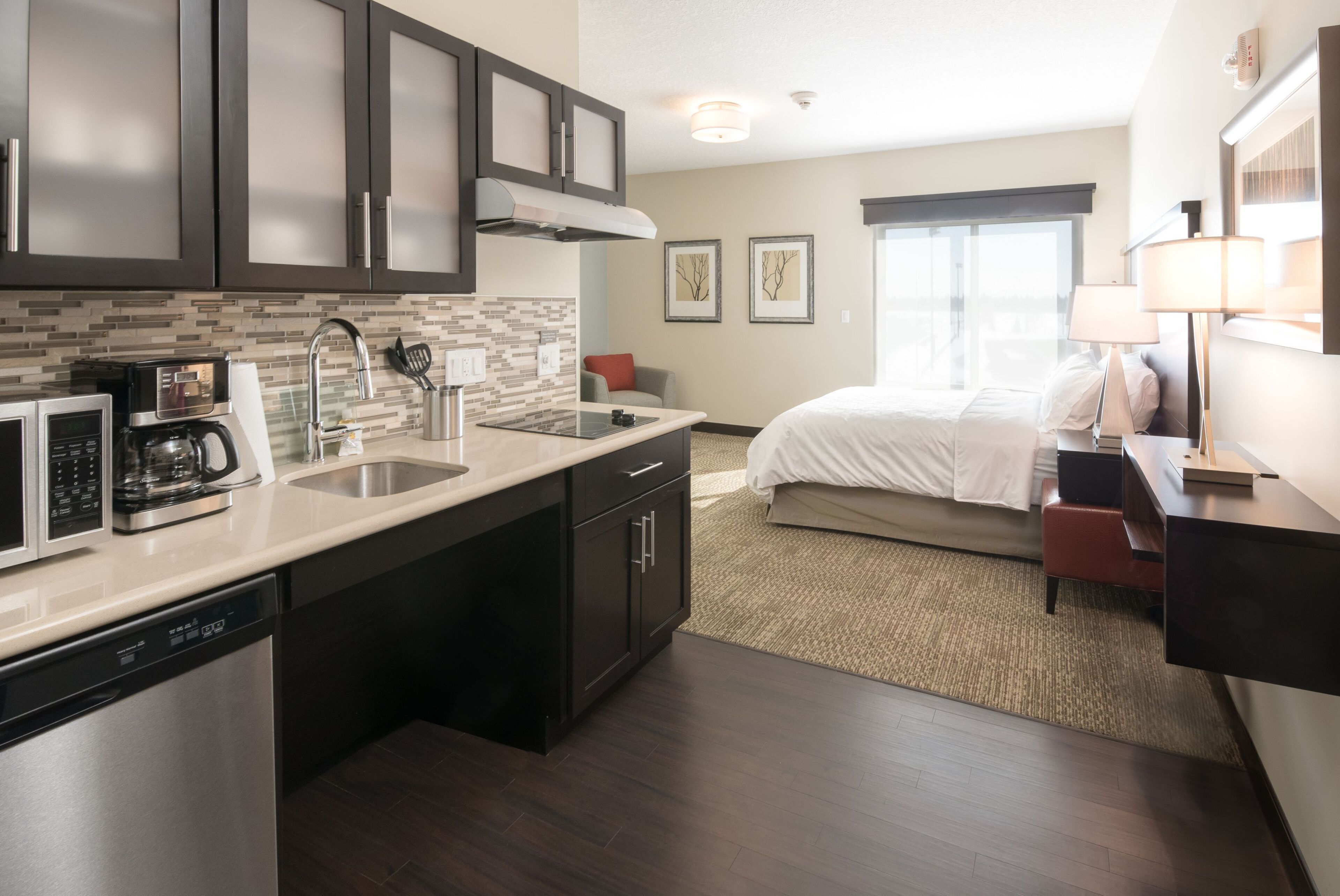 A modern kitchen with a dishwasher, microwave, and coffee maker. There is a bed with white bedding and a red chair in the adjacent bedroom.