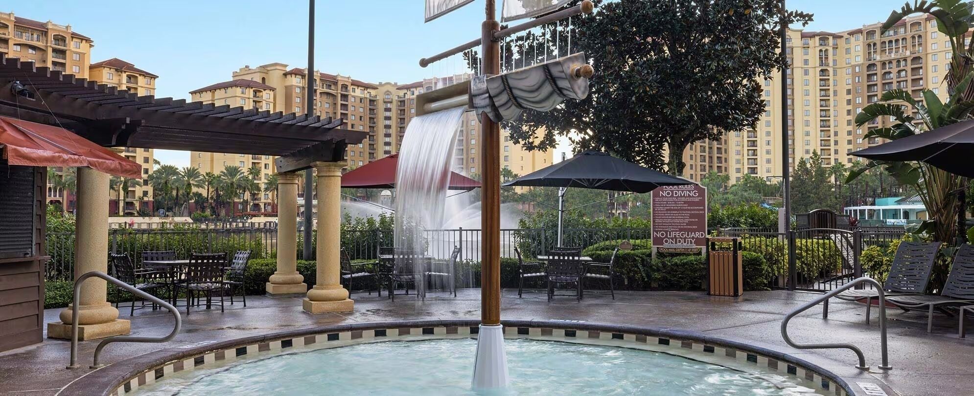 Zona de piscina de hotel con una fuente de agua, mobiliario exterior y un cartel que indica que no se permite bucear ni hay socorrista de guardia.