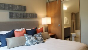 Una habitación de hotel con cama, almohadas, mesita de noche, lámpara y una puerta corredera al baño.