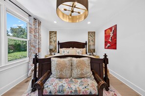 Quarto com cama de madeira, almofadas com padrão floral, luminária redonda no teto e vista para o verde lá fora.