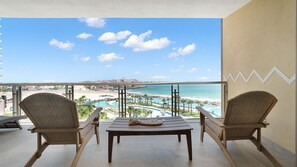 A balcony with two wicker chairs and a wooden table, overlooking a beach and a resort pool. - Two 501: Beachfront Paradise | Sonoran Star Resort (Puerto Peñasco)