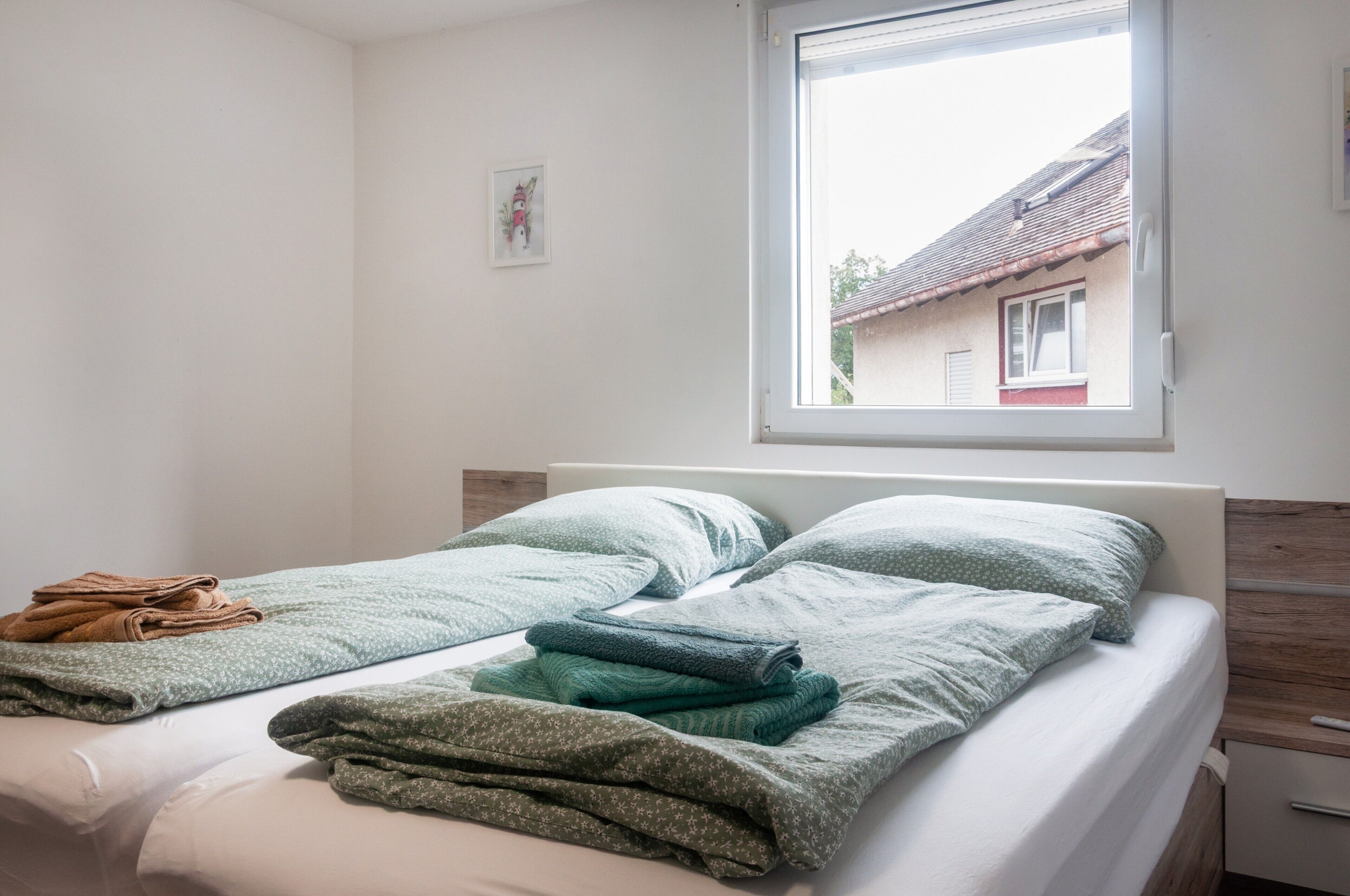 Ein Einzelbett mit Kopfteil, ein Fenster mit Blick auf ein Gebäude und ein kleiner Nachttisch mit Lampe.