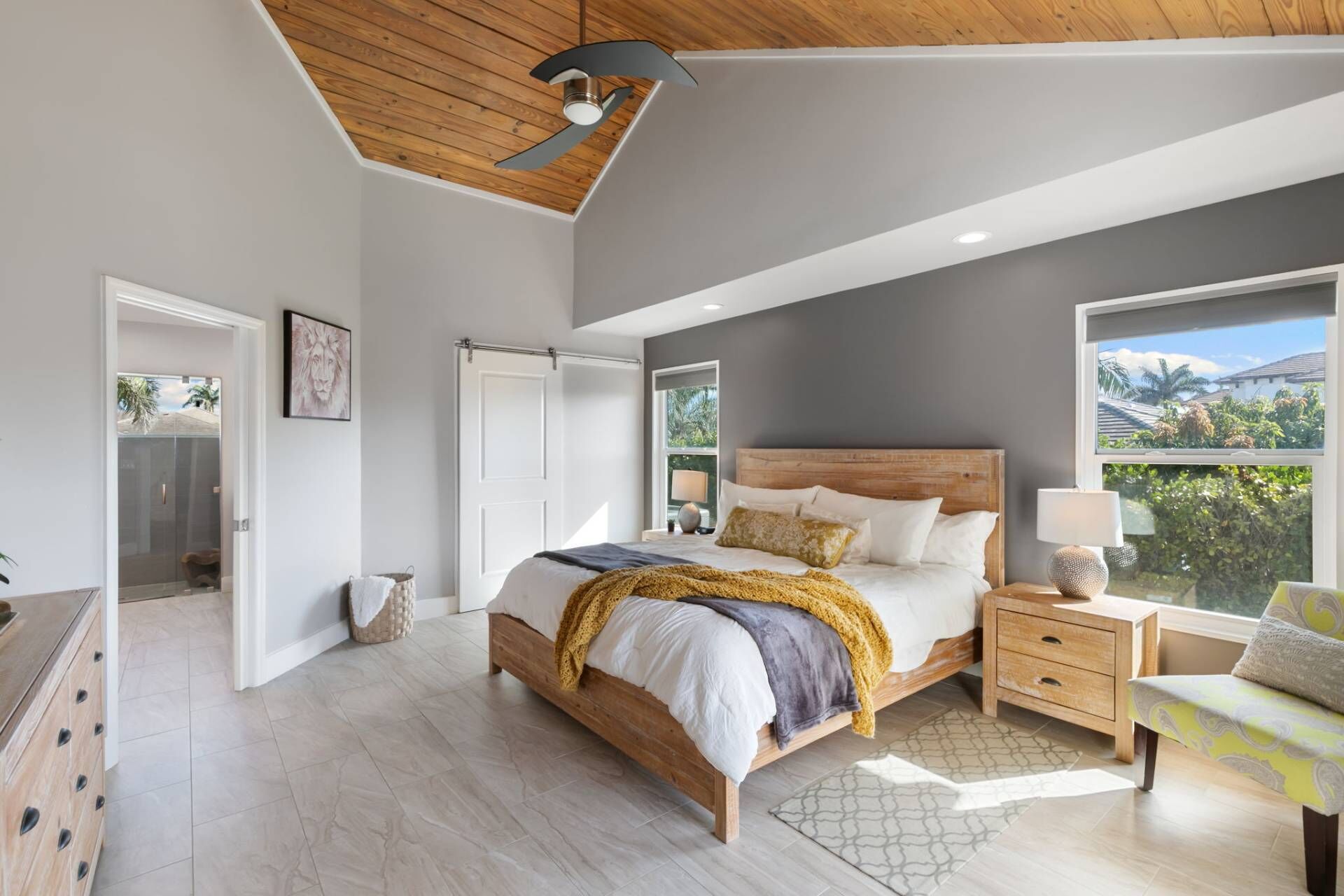 Une chambre moderne avec un plafond en bois, un grand lit avec une tête de lit, une table de chevet avec une lampe, et une vue sur la verdure à l’extérieur.