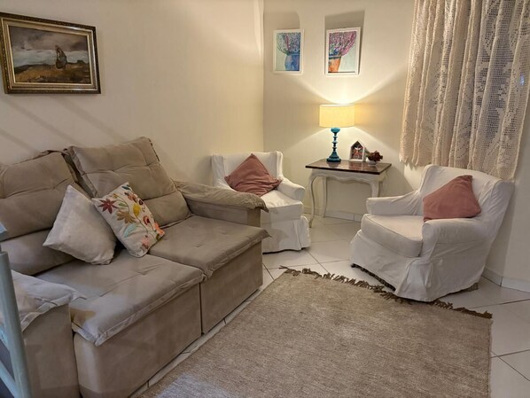A living room with a beige sofa, two white armchairs, a small table with a lamp, and a decorative rug.