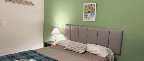 Una cama bien tendida con una colcha a cuadros, una almohada y una manta. Hay una lámpara en una mesita de noche y un cuadro en la pared.